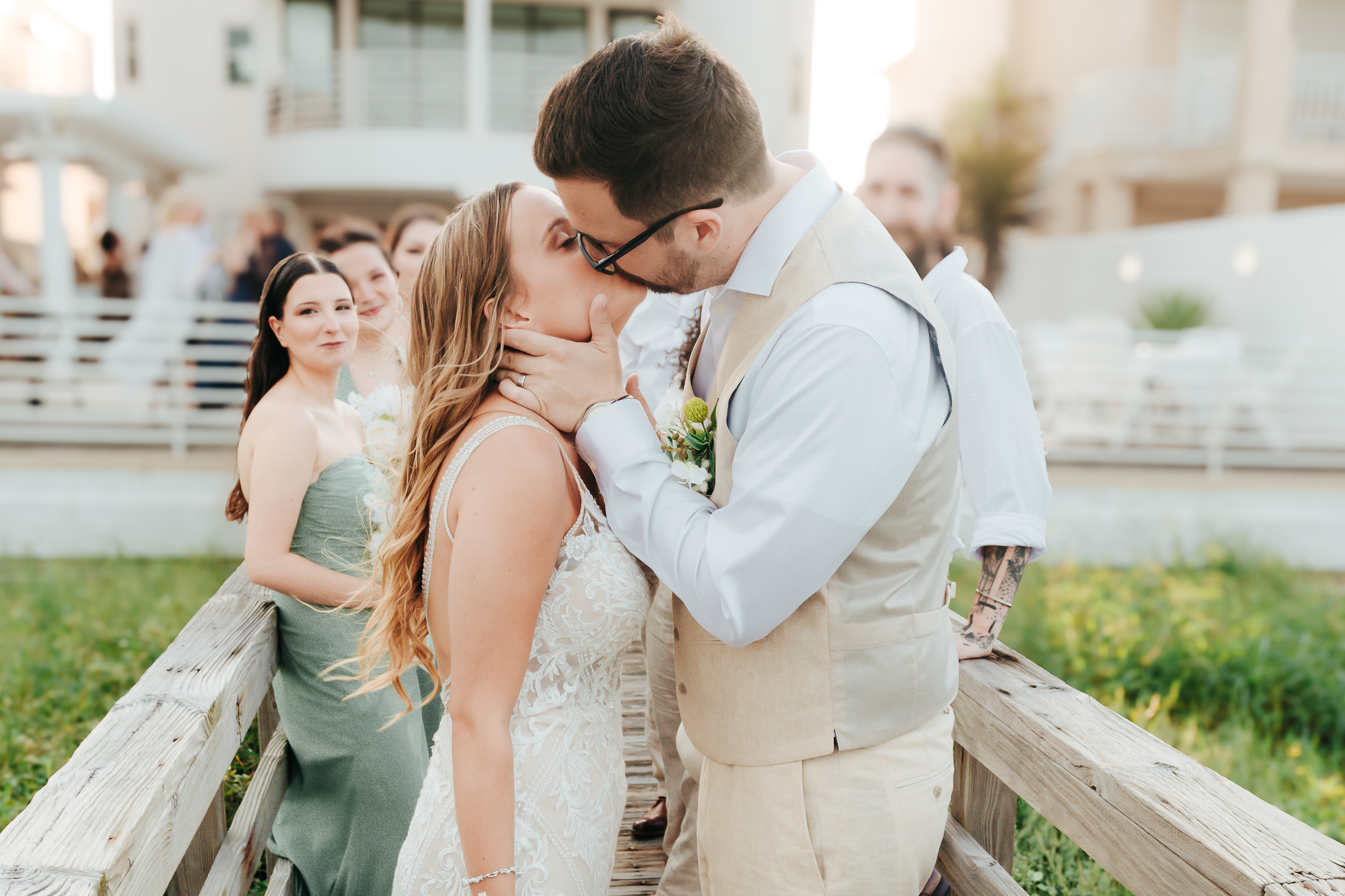 Bride and groom sharing their first kiss on a wooden boardwalk at golden hour while bridesmaids watch and smile, photographed by Weddings by Wendi