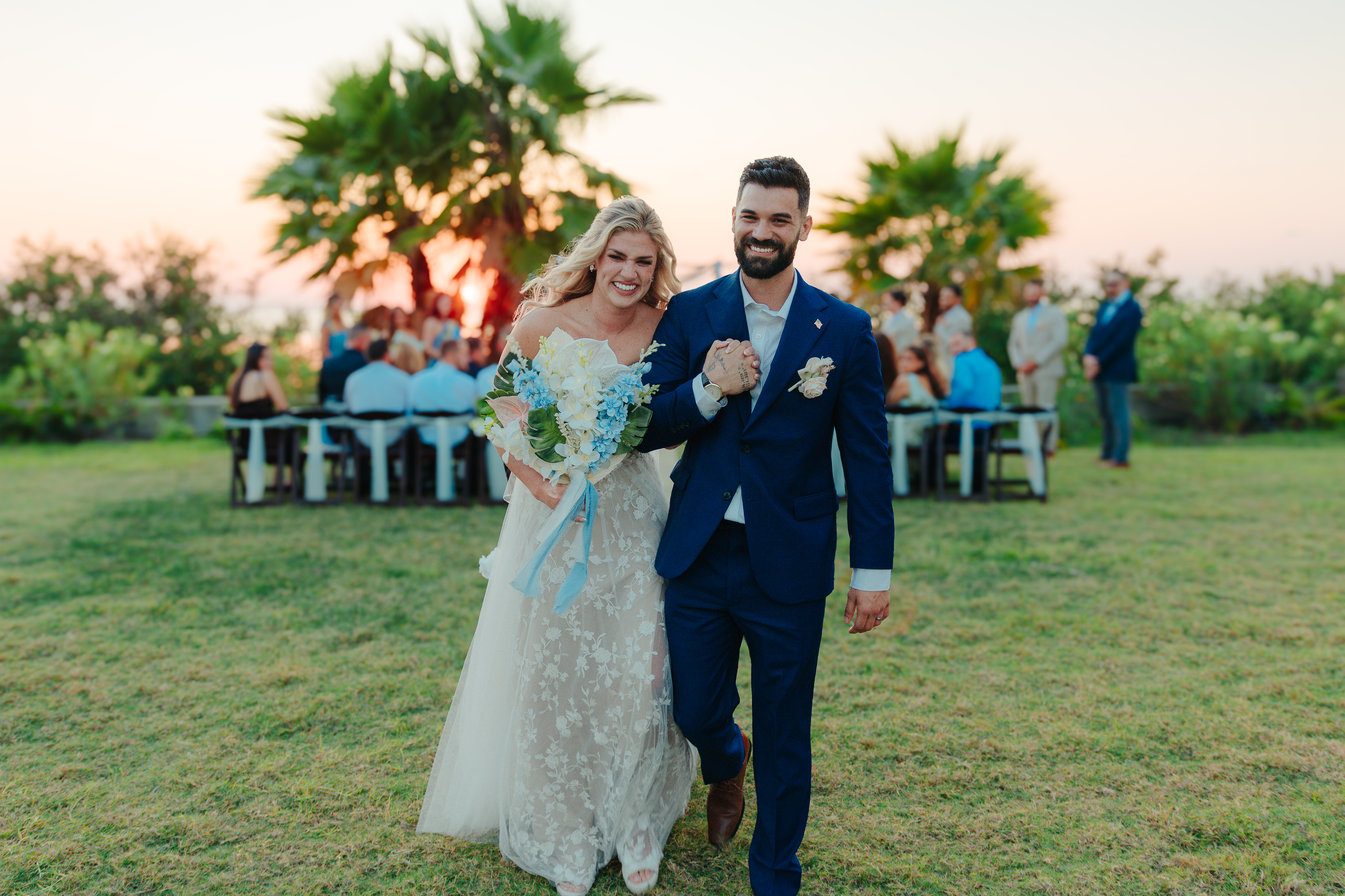 Bride and groom walking hand in hand after their ceremony at sunset, palm trees and guests in the background, photographed by Weddings by Wendi