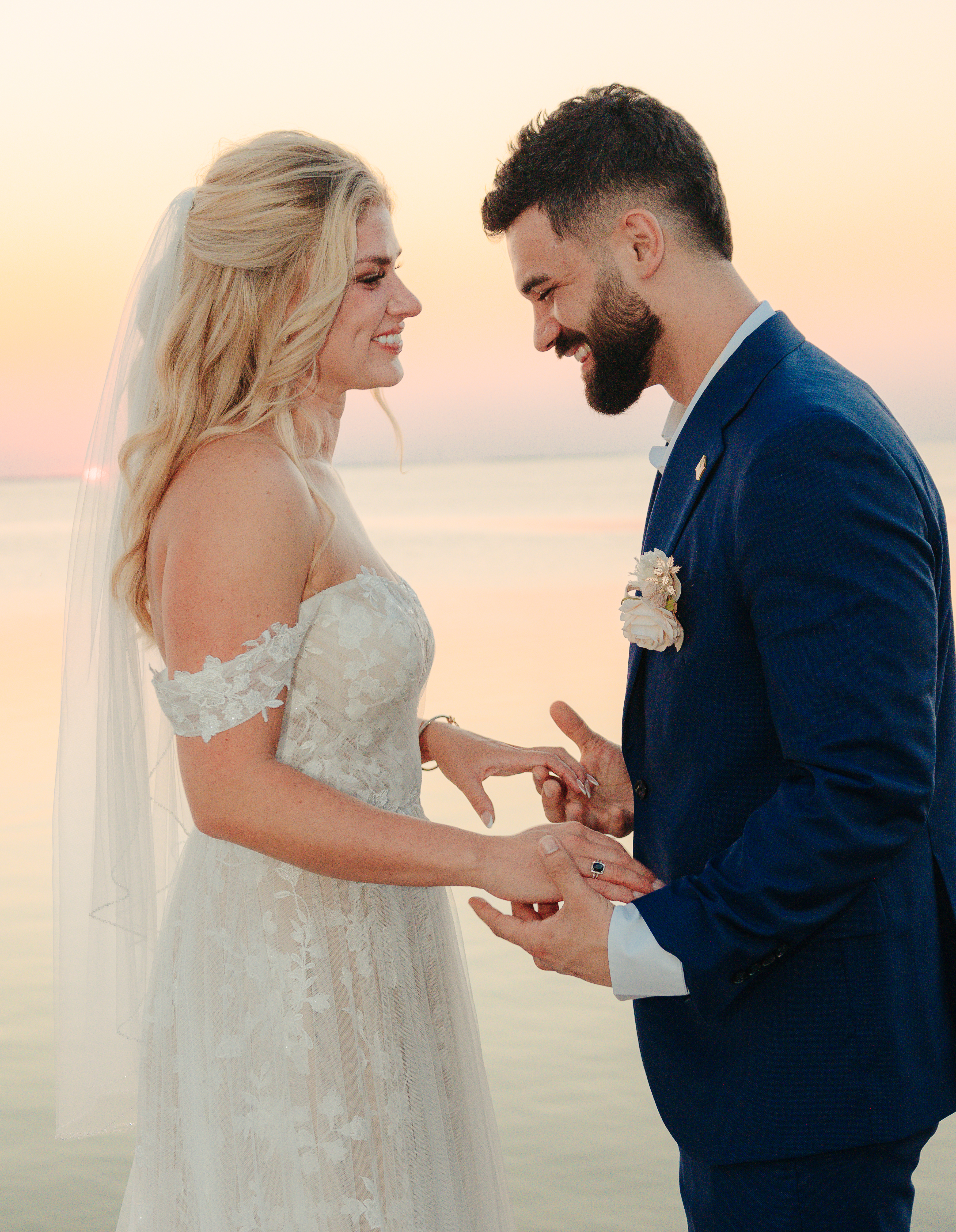 Bride and groom exchanging rings at sunset, smiling at each other with a warm golden sky over the water, photographed by Weddings by Wendi
