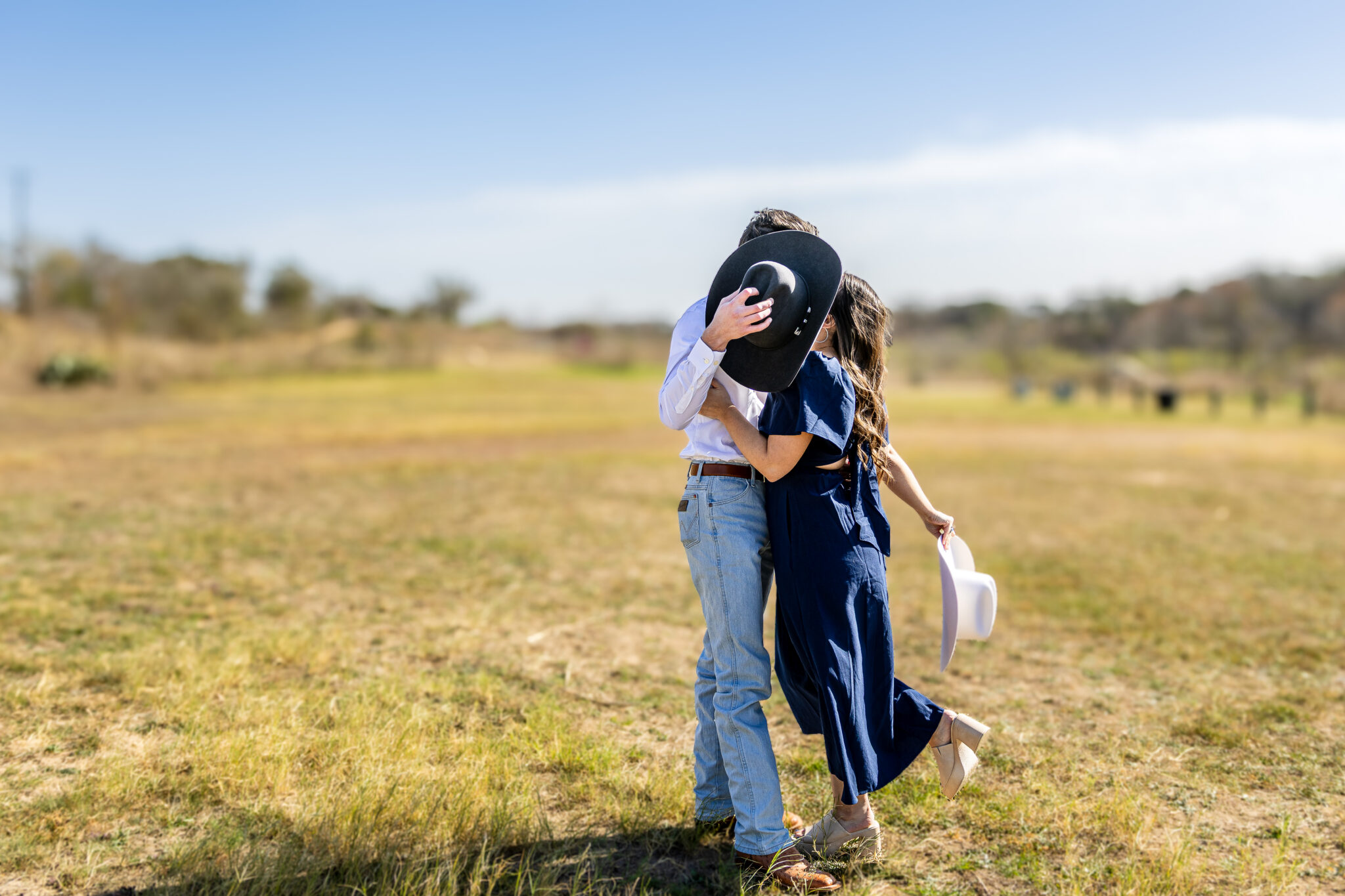 Engaged couple sharing a kiss in an open Texas Hill Country field, the groom holding a black cowboy hat over their faces, photographed by Weddings by Wendi