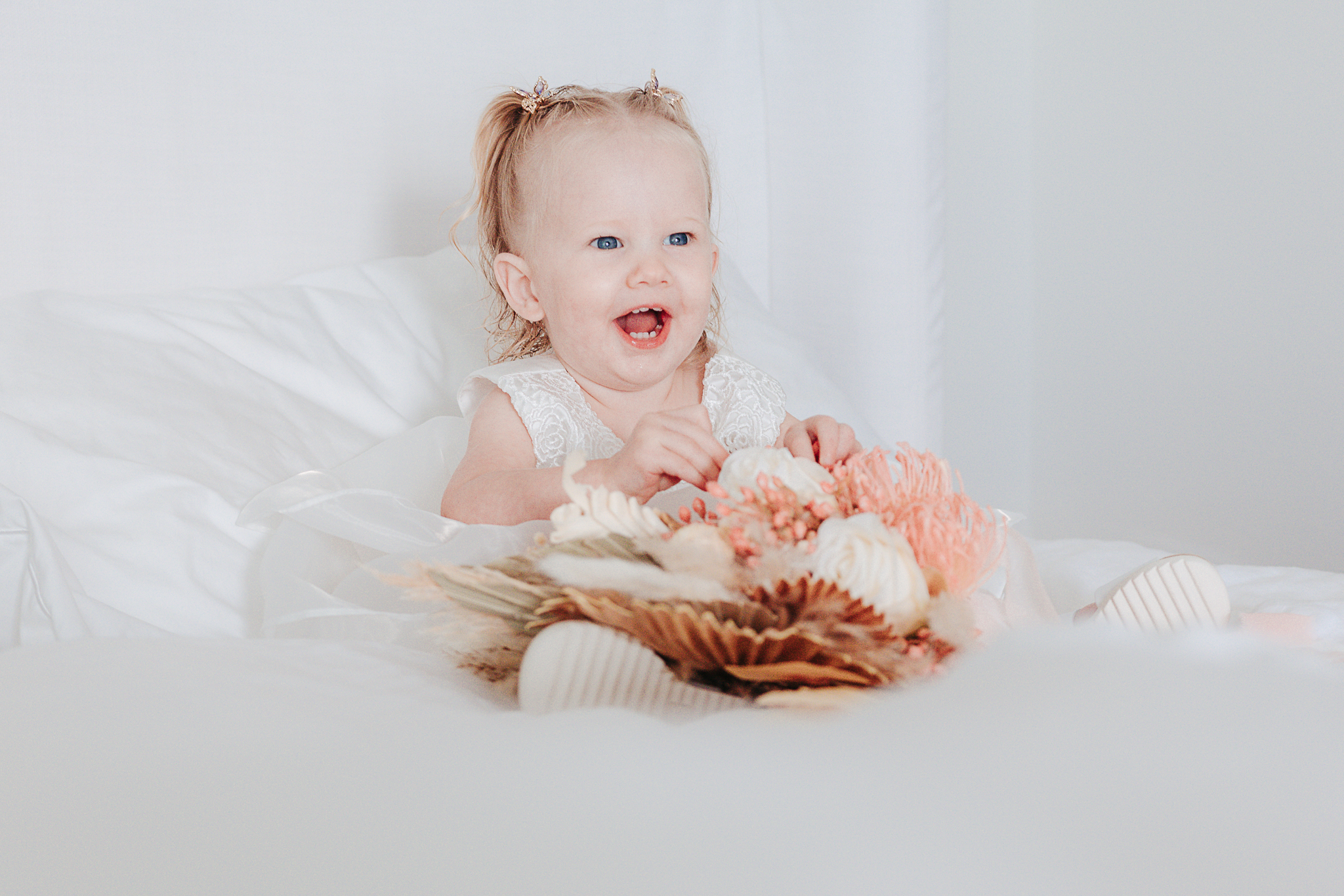 Smiling baby flower girl in a white lace dress holding a blush and dried floral bouquet, photographed by Weddings by Wendi