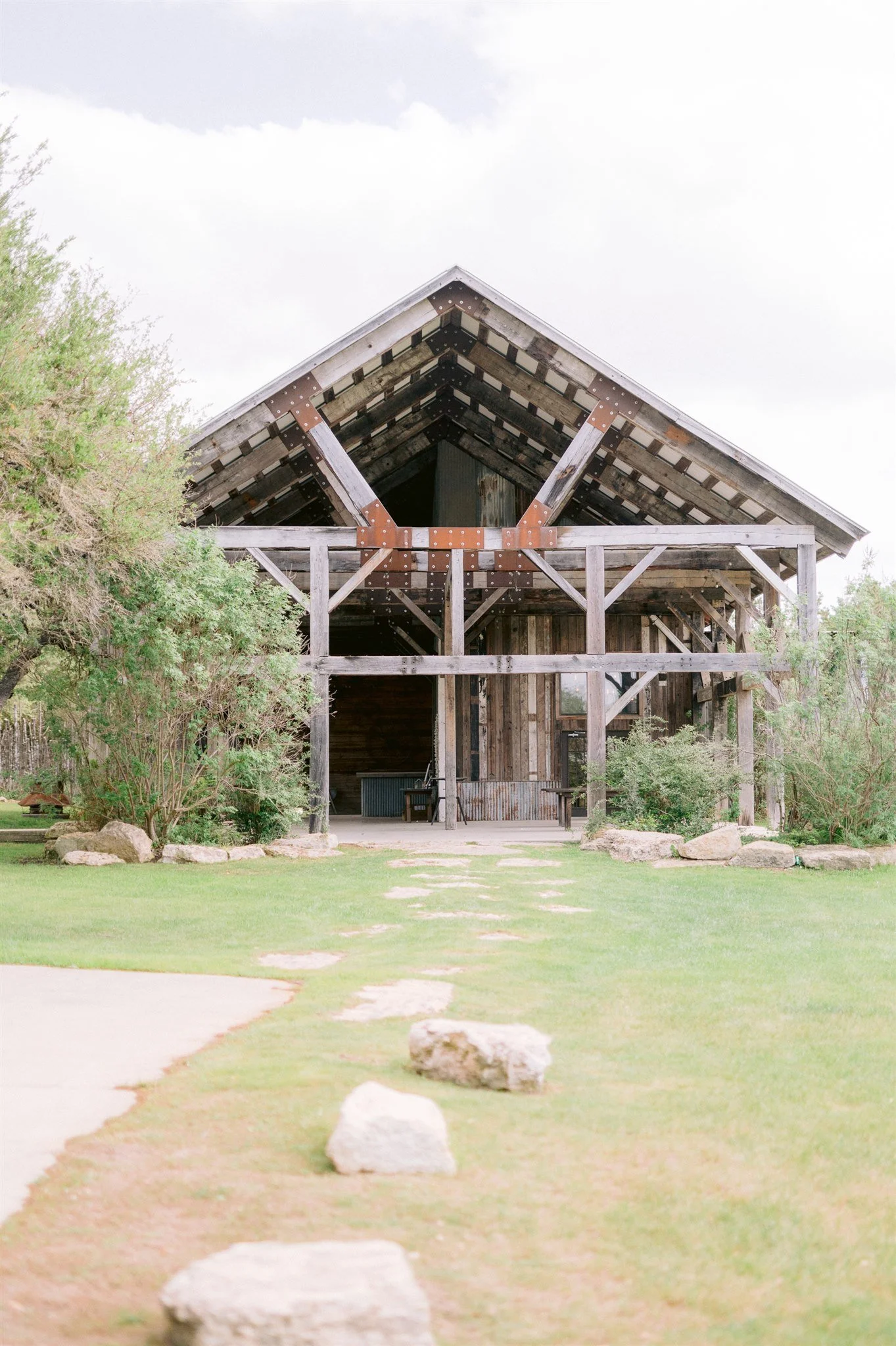 The Creek Haus rustic open-air barn exterior with reclaimed wood gable, steel cross detail, and stone pathway leading through manicured grounds in Dripping Springs Texas