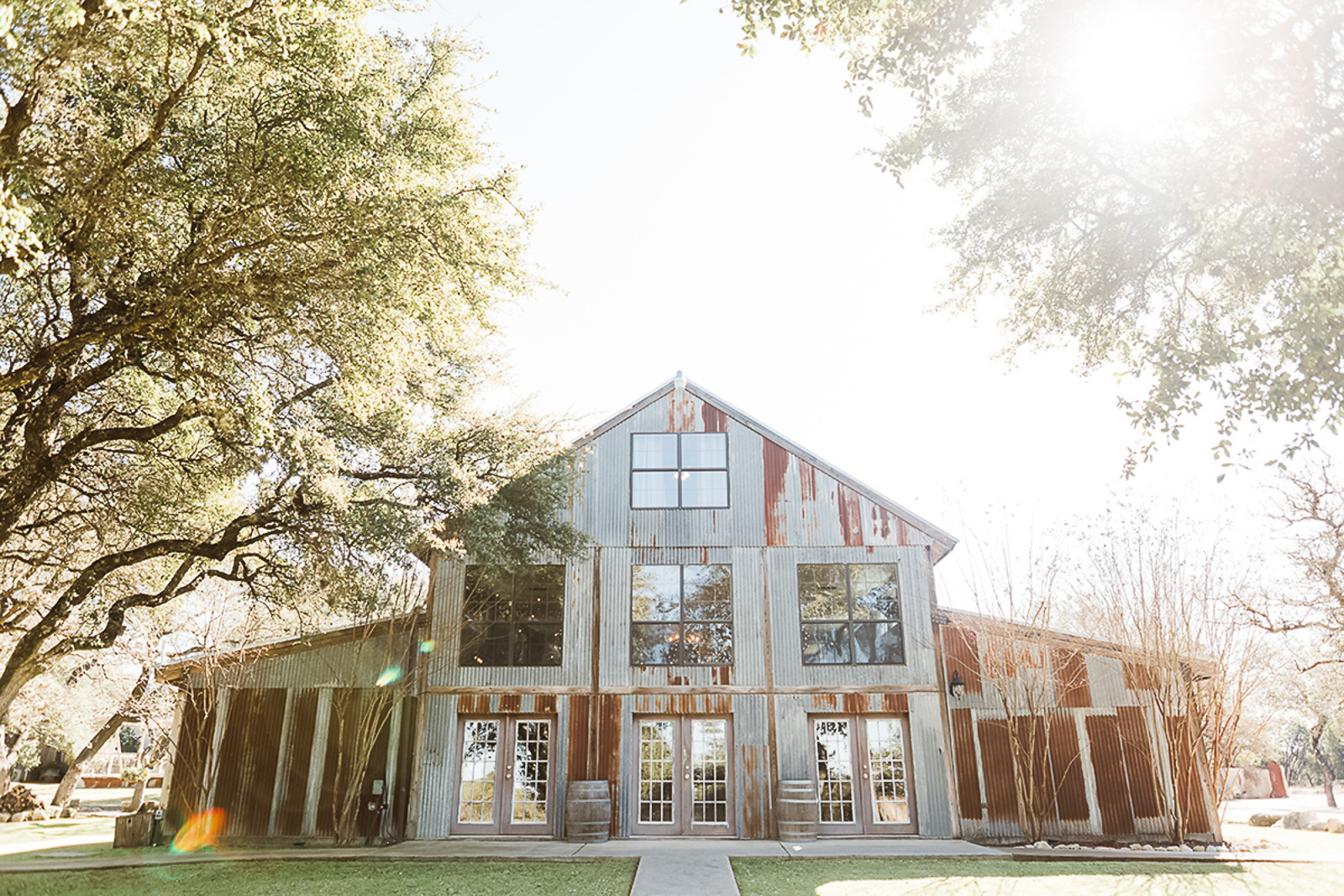 Vista West Ranch rustic corrugated metal barn facade with French doors and wine barrels, surrounded by live oak trees in Dripping Springs Texas