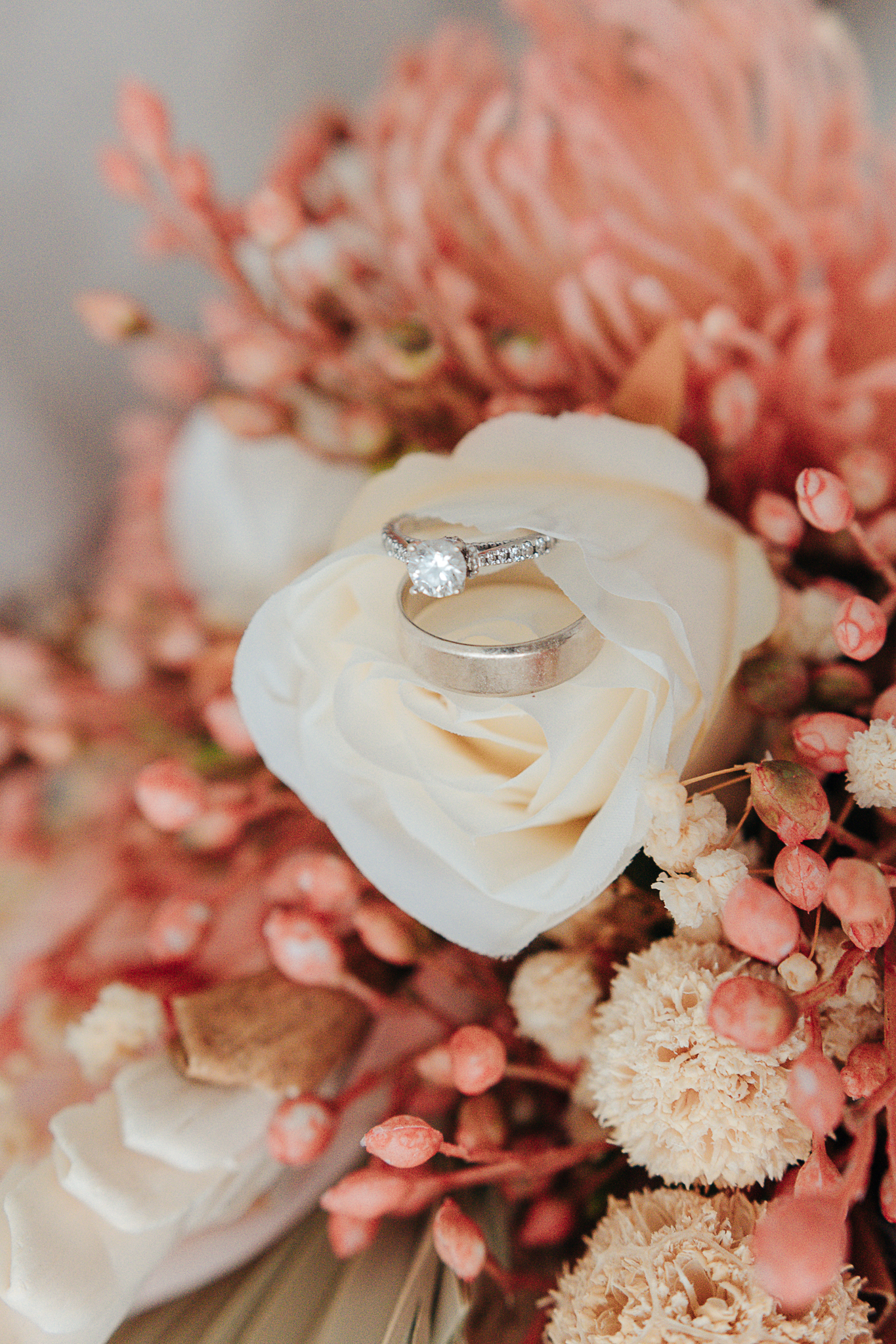 Wedding rings resting on a white rose surrounded by a blush and pink bridal bouquet, photographed by Weddings by Wendi