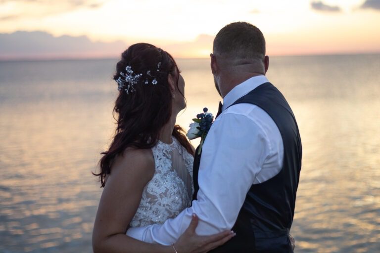 a bride and groom standing on the beach at sunset