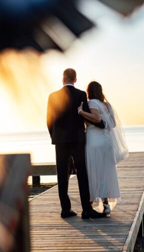 a bride and groom standing on a dock at sunset