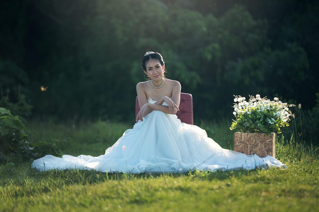 Bride in a flowing white gown sitting gracefully in a lush green field, radiating joy and elegance amidst nature's beauty.