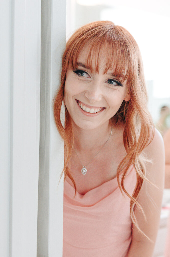 Full Wedding Planning - Weddings By Wendi Smiling woman in pink dress peeking around a doorframe, radiating joy and excitement before her wedding day.